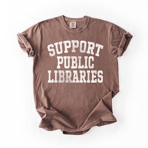 May include: A brown t-shirt with the text "SUPPORT PUBLIC LIBRARIES" in white, distressed lettering. The shirt is folded and laid flat on a white surface. The t-shirt has a crew neck and short sleeves.