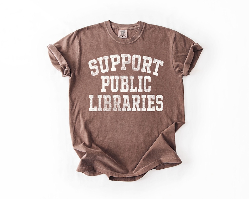 May include: A brown t-shirt with the text "SUPPORT PUBLIC LIBRARIES" in white, distressed lettering. The shirt is folded and laid flat on a white surface. The t-shirt has a crew neck and short sleeves.