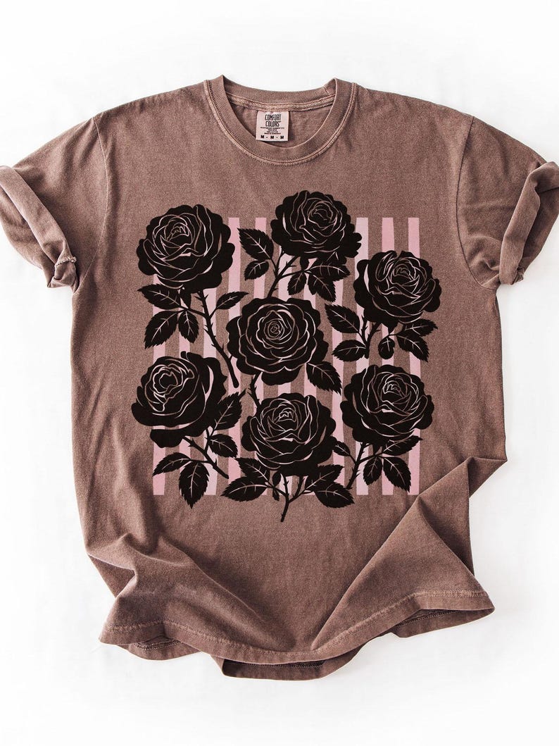 May include: A brown t-shirt featuring a black rose graphic with pink vertical stripes. The roses are detailed with leaves and stems. The shirt has a crew neck and short sleeves.