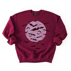 May include: A maroon sweatshirt with a distressed graphic of a full moon and bats. The bats are black, and the moon is a faded purple. The sweatshirt has long sleeves and a crew neck. A Halloween-themed garment.