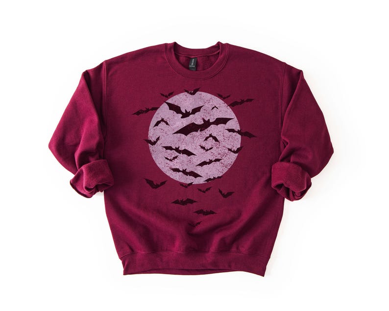 May include: A maroon sweatshirt with a distressed graphic of a full moon and bats. The bats are black, and the moon is a faded purple. The sweatshirt has long sleeves and a crew neck. A Halloween-themed garment.