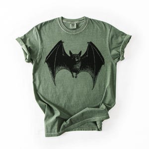 May include: A sage green t-shirt featuring a detailed black illustration of a bat with outstretched wings. The bat is centered on the shirt, with the wings spanning across the chest. The shirt has a classic crew neck and short sleeves.