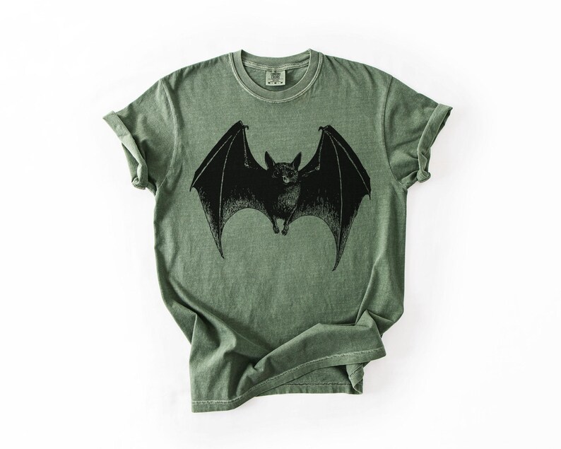 May include: A sage green t-shirt featuring a detailed black illustration of a bat with outstretched wings. The bat is centered on the shirt, with the wings spanning across the chest. The shirt has a classic crew neck and short sleeves.