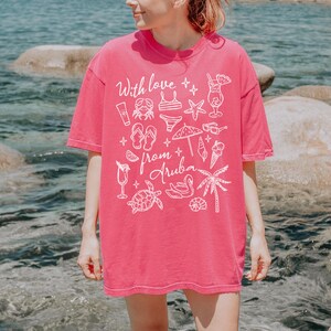 Puede incluir: Una camiseta rosa vibrante con el texto "With love from Aruba" y varias ilustraciones blancas con tem&aacute;tica playera. El dise&ntilde;o incluye un cangrejo, un bikini, gafas de sol y una palmera. La camiseta es de estilo c&oacute;modo e informal.