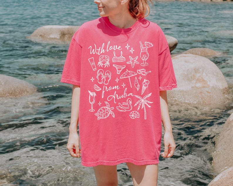 Puede incluir: Una camiseta rosa vibrante con el texto "With love from Aruba" y varias ilustraciones blancas con tem&aacute;tica playera. El dise&ntilde;o incluye un cangrejo, un bikini, gafas de sol y una palmera. La camiseta es de estilo c&oacute;modo e informal.