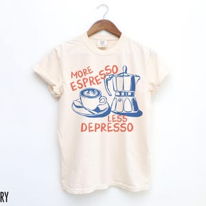 May include: A cream colored t-shirt with a blue and red graphic of a coffee pot and a cup of coffee. The text on the shirt reads "More Espresso Less Depresso".