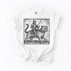 Puede incluir: Camiseta blanca con una ilustraci&oacute;n en blanco y negro de un caballero montado en un gato, sosteniendo una lanza y un escudo. El gr&aacute;fico est&aacute; enmarcado dentro de un rect&aacute;ngulo. La camiseta tiene cuello redondo y mangas cortas.