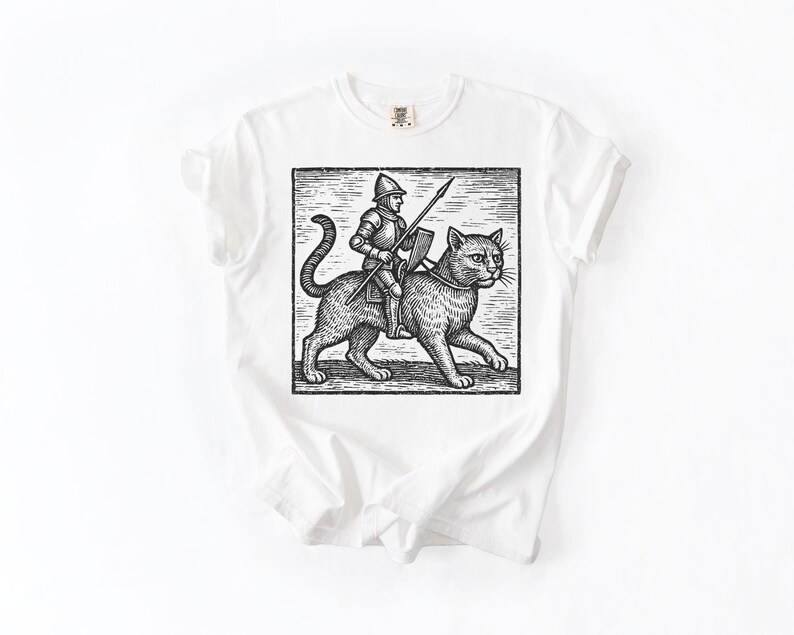 Puede incluir: Camiseta blanca con una ilustraci&oacute;n en blanco y negro de un caballero montado en un gato, sosteniendo una lanza y un escudo. El gr&aacute;fico est&aacute; enmarcado dentro de un rect&aacute;ngulo. La camiseta tiene cuello redondo y mangas cortas.