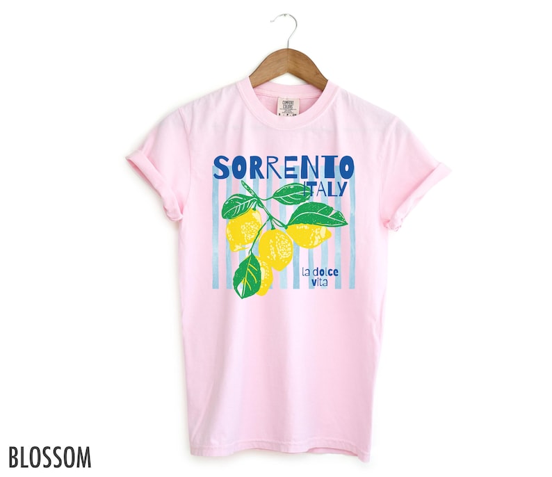 Puede incluir: Camiseta rosa claro con un dise&ntilde;o de limones y la frase "Sorrento Italy" en azul. La frase "la dolce vita" tambi&eacute;n est&aacute; presente. La palabra "Blossom" est&aacute; impresa en la esquina inferior izquierda.