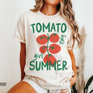 May include: A cream-colored t-shirt with a graphic of a tomato plant with the text "Tomato girl summer Italy" printed on it.