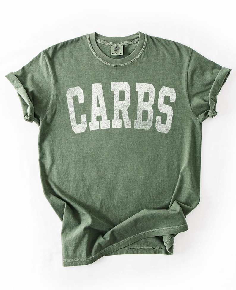 May include: Olive green t-shirt with the word "CARBS" printed in large, distressed white letters. The shirt has a crew neck and rolled-up sleeves. The fabric appears soft and slightly faded, giving it a vintage look.