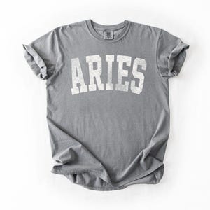 May include: A gray t-shirt with the word "ARIES" printed in white, using a distressed, collegiate-style font. The shirt is folded and has rolled-up sleeves. The t-shirt is made of a soft material.