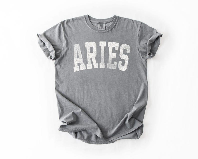 May include: A gray t-shirt with the word "ARIES" printed in white, using a distressed, collegiate-style font. The shirt is folded and has rolled-up sleeves. The t-shirt is made of a soft material.