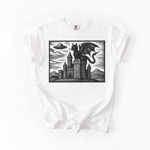 May include: White t-shirt featuring a black and white graphic of a castle with a winged cat on top. A UFO hovers in the sky above the castle. The design is a linocut style illustration.