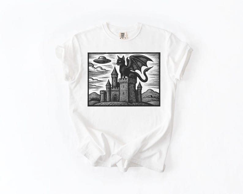 May include: White t-shirt featuring a black and white graphic of a castle with a winged cat on top. A UFO hovers in the sky above the castle. The design is a linocut style illustration.