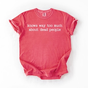 Puede incluir: Una camiseta de color coral con la frase "knows way too much about dead people" impresa en blanco. La camiseta tiene mangas cortas y cuello redondo. La tela parece suave y ligeramente descolorida.