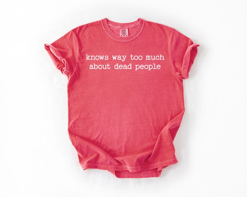 Puede incluir: Una camiseta de color coral con la frase "knows way too much about dead people" impresa en blanco. La camiseta tiene mangas cortas y cuello redondo. La tela parece suave y ligeramente descolorida.