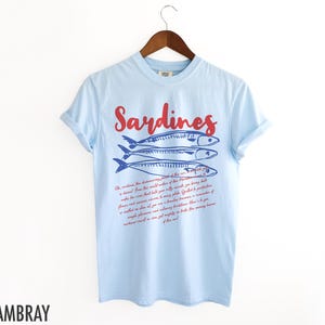May include: Light blue t-shirt with a red and blue graphic of sardines and a text description about sardines. The text reads "Oh, sardines! The shimmering jewel of the sea. No creature from the world's waters of the Mediterranean is a distant, cold, and distant relative to this sardine. It is a treasure from that warm shore, perfectly preserved, full of oceanic flavor and a timeless treasure. Grilled, it is a realted phenomenon and incredibly flavorful. Here is your sardine, and all its velvety traditions. The many treasures of the sea."