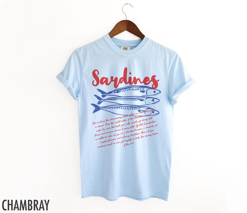 May include: Light blue t-shirt with a red and blue graphic of sardines and a text description about sardines. The text reads "Oh, sardines! The shimmering jewel of the sea. No creature from the world's waters of the Mediterranean is a distant, cold, and distant relative to this sardine. It is a treasure from that warm shore, perfectly preserved, full of oceanic flavor and a timeless treasure. Grilled, it is a realted phenomenon and incredibly flavorful. Here is your sardine, and all its velvety traditions. The many treasures of the sea."