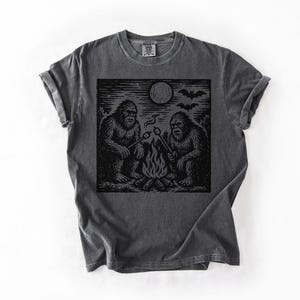 May include: Dark gray t-shirt featuring a black graphic of two Bigfoot creatures roasting marshmallows over a campfire under a full moon. Bats are flying in the sky above the moon. The shirt has a crew neck and short sleeves.