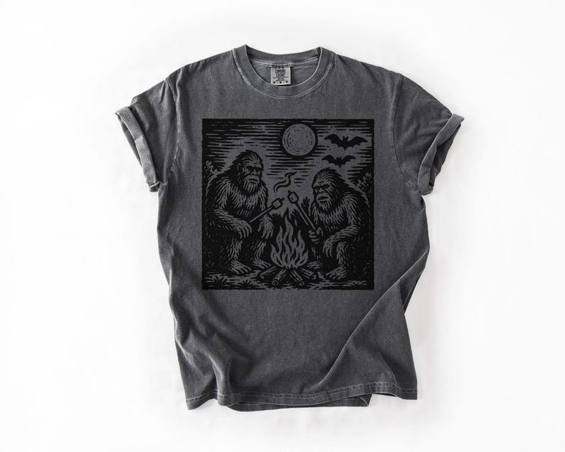 May include: Dark gray t-shirt featuring a black graphic of two Bigfoot creatures roasting marshmallows over a campfire under a full moon. Bats are flying in the sky above the moon. The shirt has a crew neck and short sleeves.