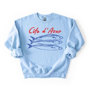 K&ouml;nnte beinhalten: Hellblaues Sweatshirt mit dem roten Text "C&ocirc;te d'Azur" &uuml;ber drei blauen Fischillustrationen. Das Sweatshirt hat lange &Auml;rmel und einen Rundhalsausschnitt. Das Design ist einfach und maritim.
