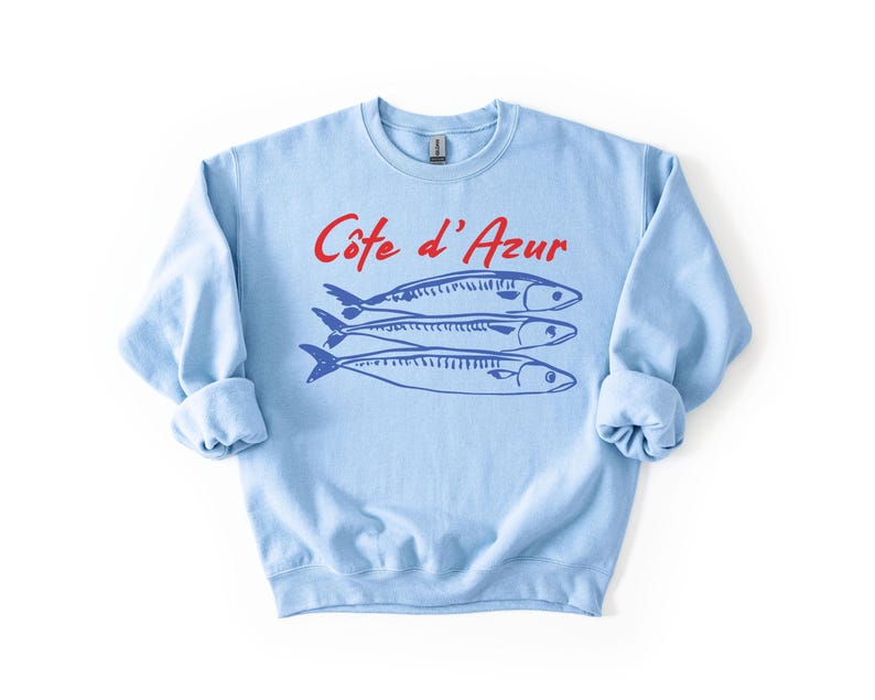 K&ouml;nnte beinhalten: Hellblaues Sweatshirt mit dem roten Text "C&ocirc;te d'Azur" &uuml;ber drei blauen Fischillustrationen. Das Sweatshirt hat lange &Auml;rmel und einen Rundhalsausschnitt. Das Design ist einfach und maritim.