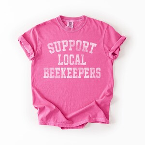 Puede incluir: Una camiseta rosa brillante con las mangas enrolladas. La camiseta presenta el texto "SUPPORT LOCAL BEEKEEPERS" en blanco, con una fuente de estilo universitario. La camiseta est&aacute; hecha de un material suave y probablemente sea de corte unisex.
