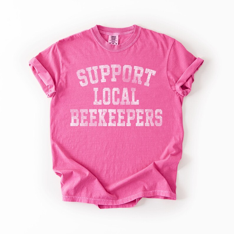 Puede incluir: Una camiseta rosa brillante con las mangas enrolladas. La camiseta presenta el texto "SUPPORT LOCAL BEEKEEPERS" en blanco, con una fuente de estilo universitario. La camiseta est&aacute; hecha de un material suave y probablemente sea de corte unisex.