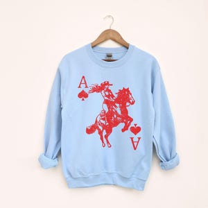 May include: Pale blue sweatshirt featuring a red graphic of a person riding a horse, with the Ace of Spades and Ace of Hearts card symbols. The sweatshirt has long sleeves and a crew neck.