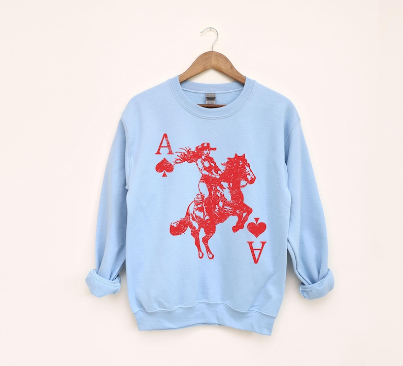 May include: Pale blue sweatshirt featuring a red graphic of a person riding a horse, with the Ace of Spades and Ace of Hearts card symbols. The sweatshirt has long sleeves and a crew neck.