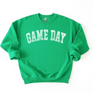 May include: A bright green sweatshirt with the text "GAME DAY" in distressed white lettering across the chest. The sweatshirt has a crew neck and long sleeves with rolled cuffs. The fabric appears soft and comfortable.