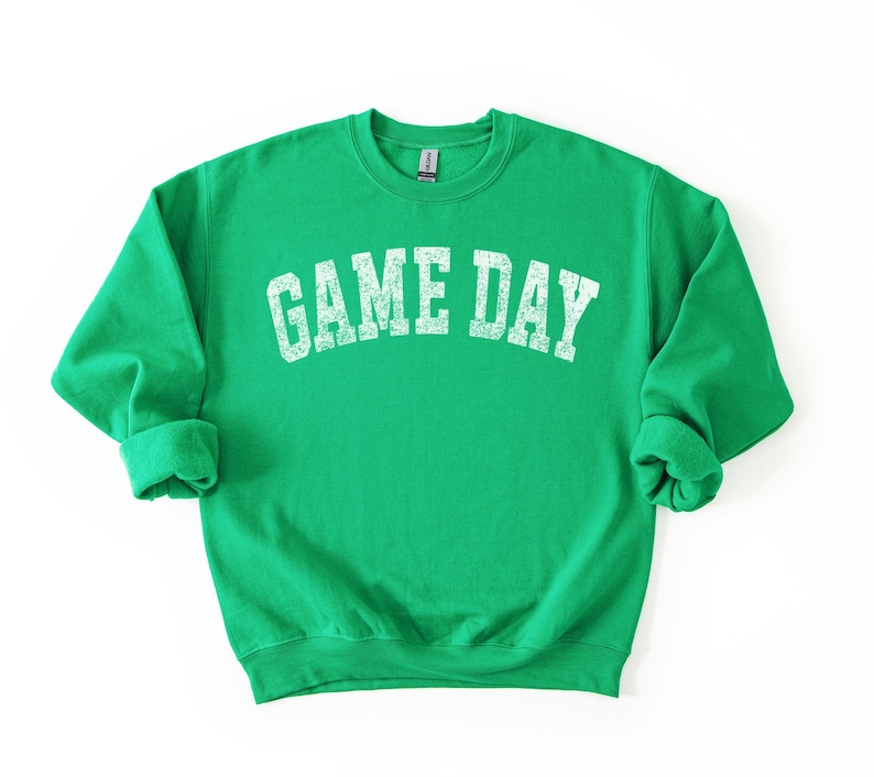 May include: A bright green sweatshirt with the text "GAME DAY" in distressed white lettering across the chest. The sweatshirt has a crew neck and long sleeves with rolled cuffs. The fabric appears soft and comfortable.