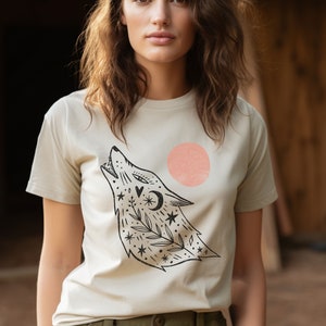 May include: A beige t-shirt featuring a black and white illustration of a wolf howling at the moon. The wolf is surrounded by stars and flowers. There is a pink circle behind the wolf.