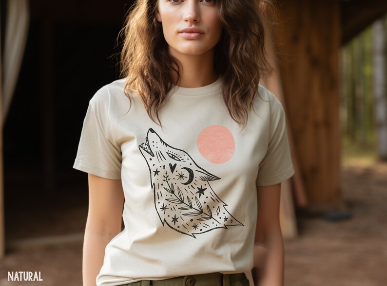 May include: A beige t-shirt featuring a black and white illustration of a wolf howling at the moon. The wolf is surrounded by stars and flowers. There is a pink circle behind the wolf.