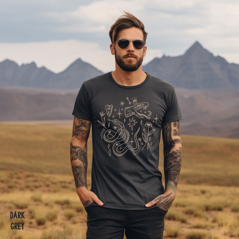 Puede incluir: Una camiseta gris oscuro con un dise&ntilde;o gr&aacute;fico de una serpiente, una corona, un diente, una estrella y otros s&iacute;mbolos. El dise&ntilde;o es de color dorado claro.