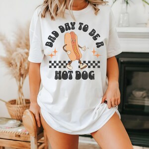 May include: White t-shirt with a graphic of a hot dog with arms and legs. The hot dog is wearing a smile and is holding a mustard bottle. The text on the shirt reads "Bad Day To Be A Hot Dog".