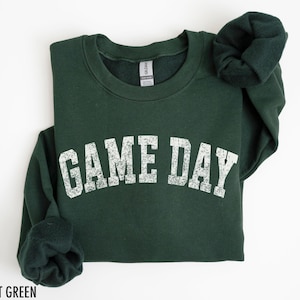 May include: Forest green sweatshirt with the text "GAME DAY" in distressed white lettering. The sweatshirt is folded, with the sleeves and collar visible. The Gildan brand tag is visible on the inside of the collar.