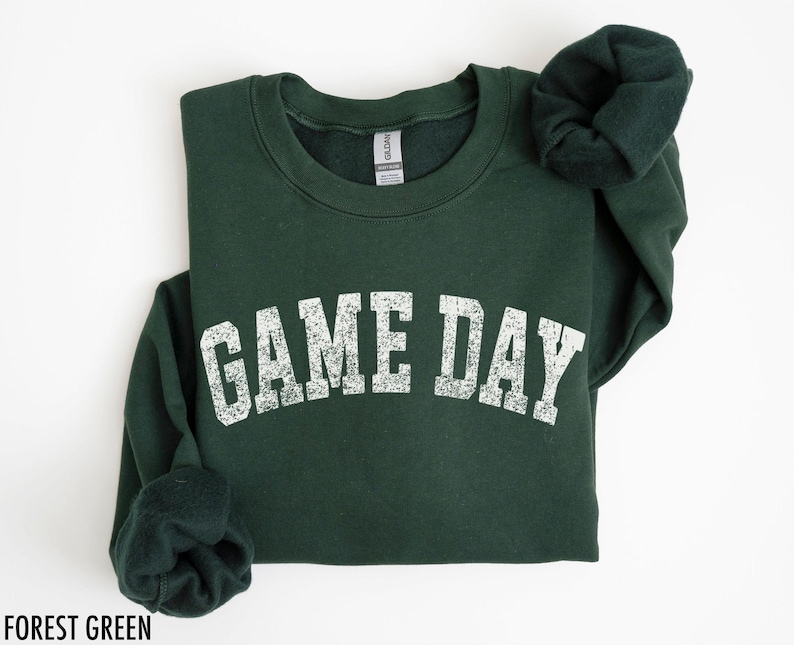 May include: Forest green sweatshirt with the text "GAME DAY" in distressed white lettering. The sweatshirt is folded, with the sleeves and collar visible. The Gildan brand tag is visible on the inside of the collar.