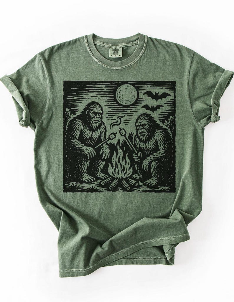 May include: Olive green t-shirt featuring a black graphic of two Bigfoot creatures roasting marshmallows over a campfire under a full moon and bats. The shirt has a crew neck and short sleeves.