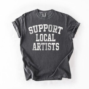 May include: Dark gray t-shirt with rolled sleeves. The shirt features the white text "SUPPORT LOCAL ARTISTS" in a collegiate font. The shirt is laid flat on a white surface.