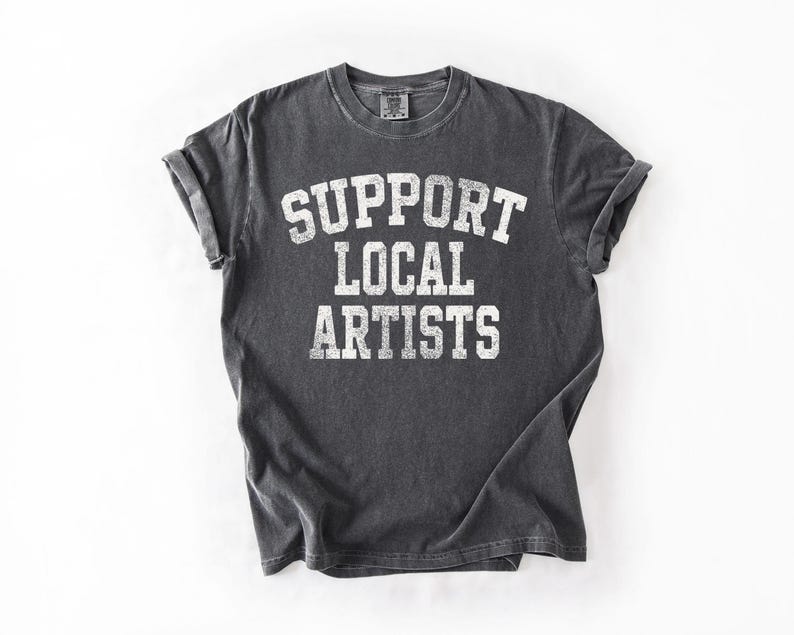 May include: Dark gray t-shirt with rolled sleeves. The shirt features the white text "SUPPORT LOCAL ARTISTS" in a collegiate font. The shirt is laid flat on a white surface.
