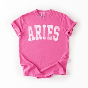 May include: A bright pink t-shirt with the word "ARIES" printed in distressed white lettering. The shirt has rolled-up sleeves and a crew neck. The fabric is slightly wrinkled, giving it a casual look.