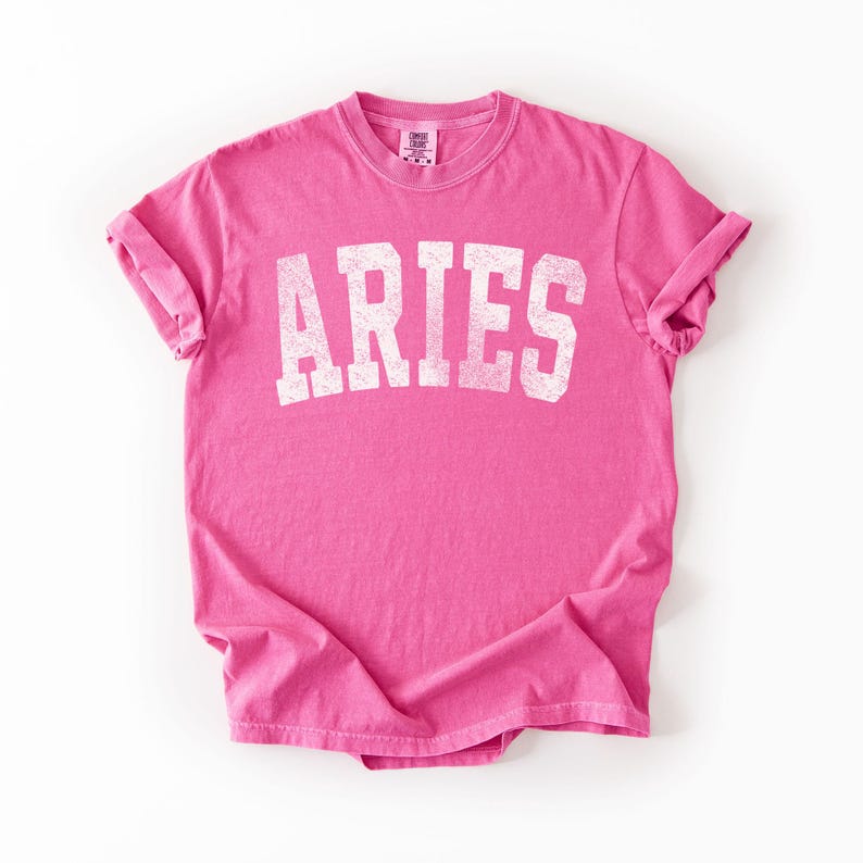 May include: A bright pink t-shirt with the word "ARIES" printed in distressed white lettering. The shirt has rolled-up sleeves and a crew neck. The fabric is slightly wrinkled, giving it a casual look.
