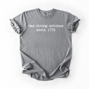 Puede incluir: Camiseta gris con el texto blanco "has strong opinions about 1776". La camiseta es de material suave y tiene las mangas enrolladas. La camiseta est&aacute; colocada sobre un fondo blanco.