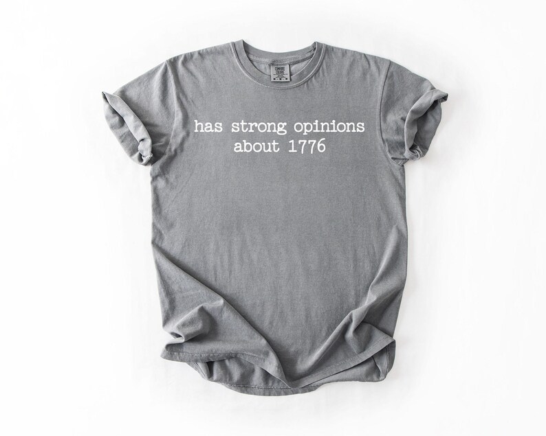 Puede incluir: Camiseta gris con el texto blanco "has strong opinions about 1776". La camiseta es de material suave y tiene las mangas enrolladas. La camiseta est&aacute; colocada sobre un fondo blanco.