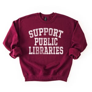 May include: A maroon sweatshirt with the words "SUPPORT PUBLIC LIBRARIES" printed in white, distressed lettering. The sweatshirt has a crew neck and long sleeves with rolled cuffs. The background is plain white.