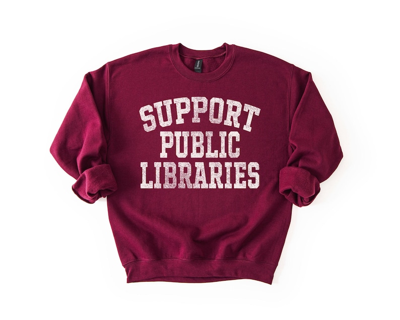 May include: A maroon sweatshirt with the words "SUPPORT PUBLIC LIBRARIES" printed in white, distressed lettering. The sweatshirt has a crew neck and long sleeves with rolled cuffs. The background is plain white.