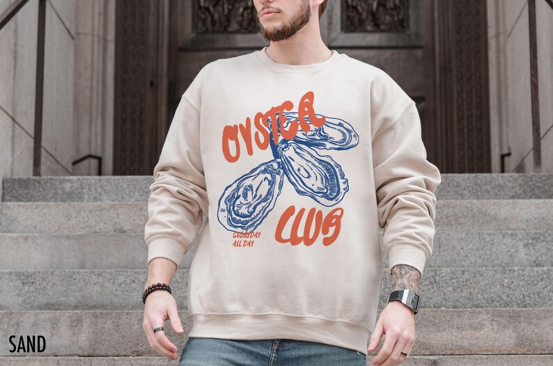 K&ouml;nnte beinhalten: Ein beiger Pullover mit einem Grafikdruck von Austern und dem Text "Oyster Club" in Orange und Blau. Der Text "Everyday All Day" ist in kleinerer Schrift unterhalb der Grafik gedruckt.