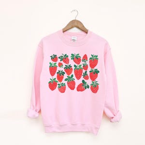 K&ouml;nnte beinhalten: Ein rosa Sweatshirt mit einem grafischen Druck von roten Erdbeeren. Die Erdbeeren sind in einem Muster auf der Vorderseite des Sweatshirts angeordnet.